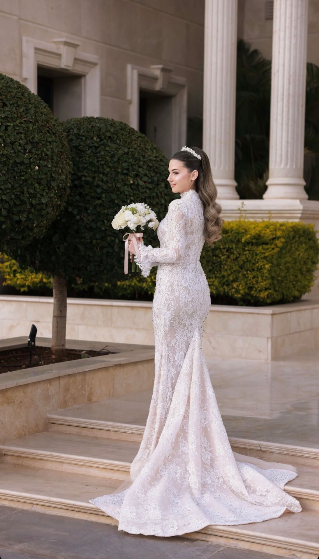 Bride in a lace gown holding a white bouquet on marble steps