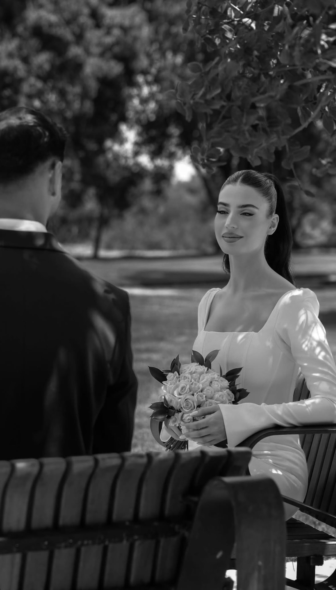 Black and white photograph of the bride seated on a park bench facing the groom