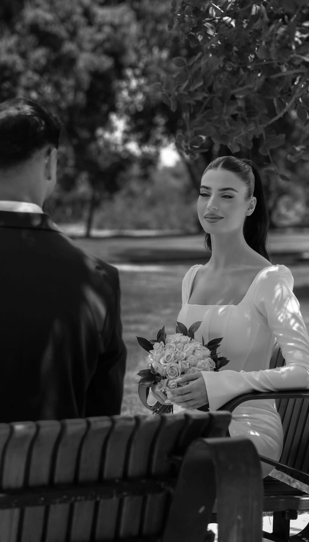 Black and white photograph of the bride seated on a park bench facing the groom