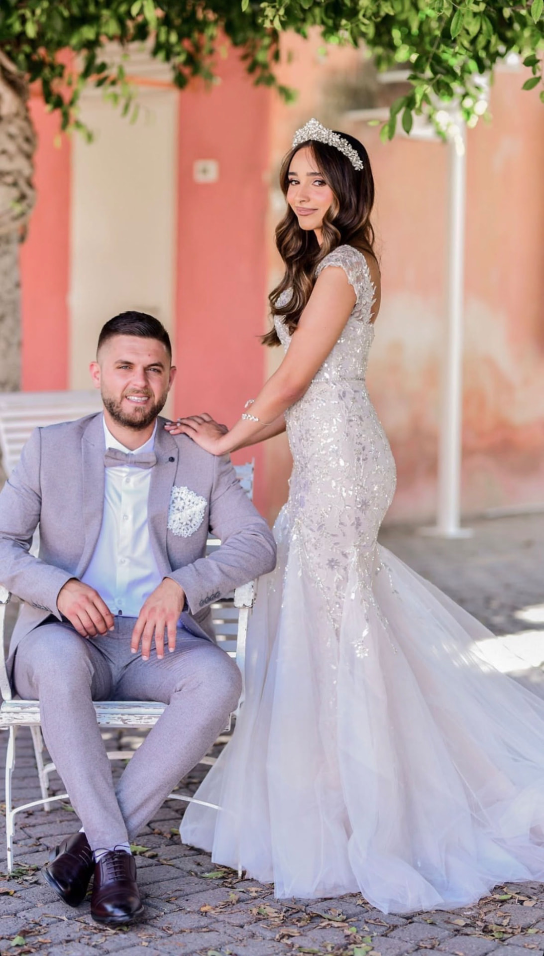 Bride wearing a tiara beside the groom in a light grey suit seated outdoors