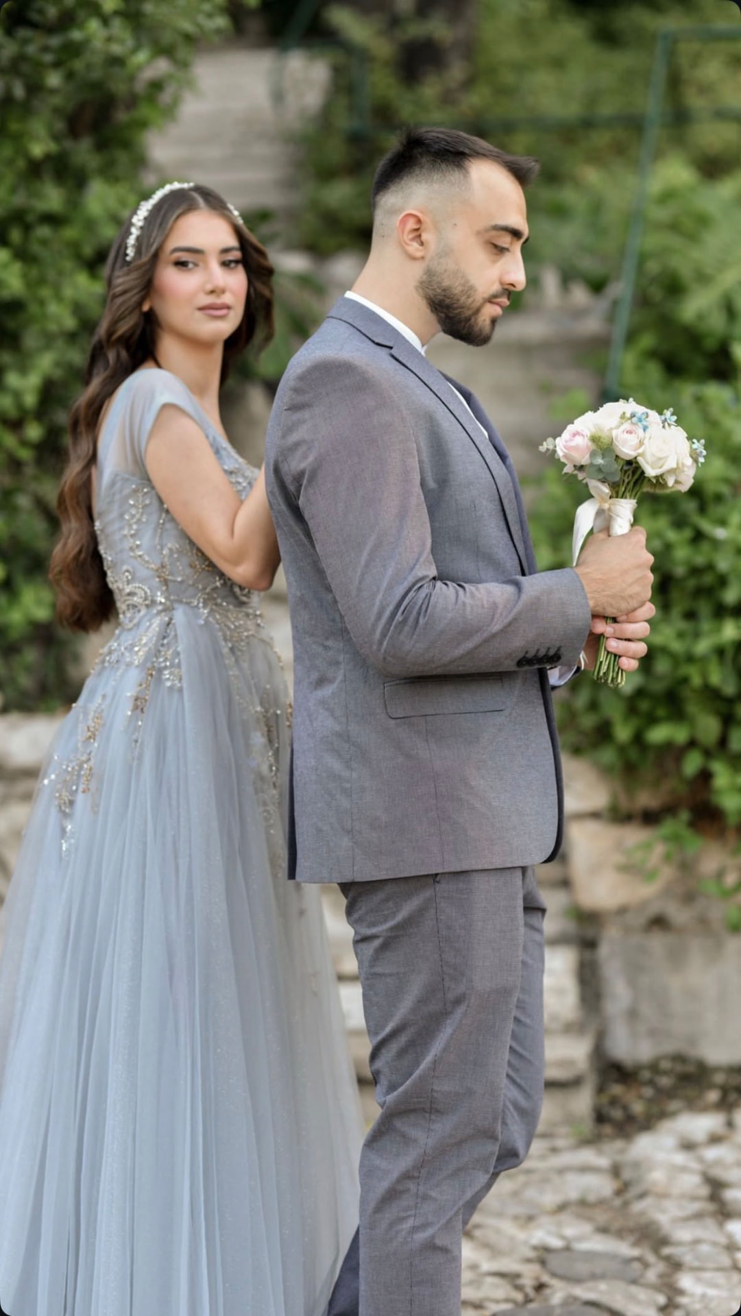 Bride in a dusty blue gown standing back to back with the groom holding a bouquet