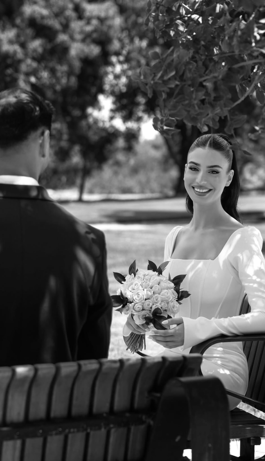 Black and white photograph of the bride smiling with her bouquet on a bench